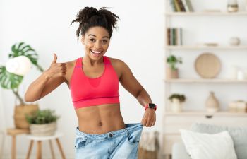 Happy Afro-American woman up in old oversize jeans presenting results of weight loss and showing her thumb up.